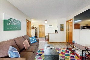 Ein Wohnzimmer mit braunem Sofa, Couchtisch und farbenfrohem Teppich. Es gibt eine Küchenzeile mit Arbeitsplatte und Mikrowelle.