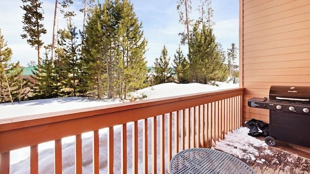Une terrasse en bois avec un barbecue et une table, entourée de neige et d’arbres.