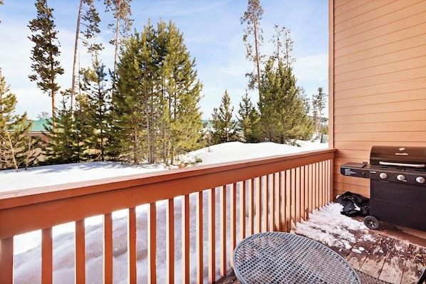 A wooden deck with a grill and a table, surrounded by snow and trees.