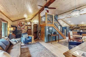 Ein großzügiger Innenraum mit Holzbalkendecke, einer Treppe zu einer oberen Ebene, einer gemütlichen Sitzecke mit Sofa und Sesseln sowie einer Küchenzeile.