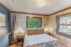 Ein Schlafzimmer einer Holzhütte mit einem Bett, einem Fenster mit Blick auf Felsen, einem Nachttisch mit Lampe und einem Wandbild.