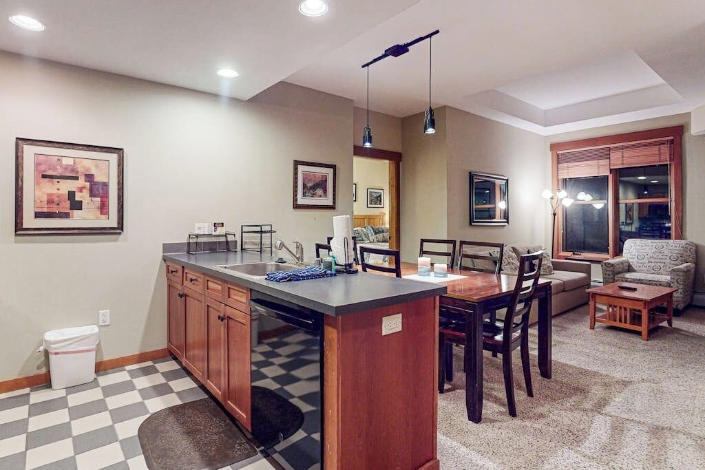 A kitchen with a central island, dining area with chairs, a sofa, and a window with blinds.