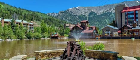 A lakeside hotel with a rustic fountain, stone benches, and mountain views.