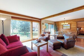 Ein geräumiges Wohnzimmer mit einem roten Sofa, Ledersesseln, einem Couchtisch und großen Fenstern mit Blick ins Freie.