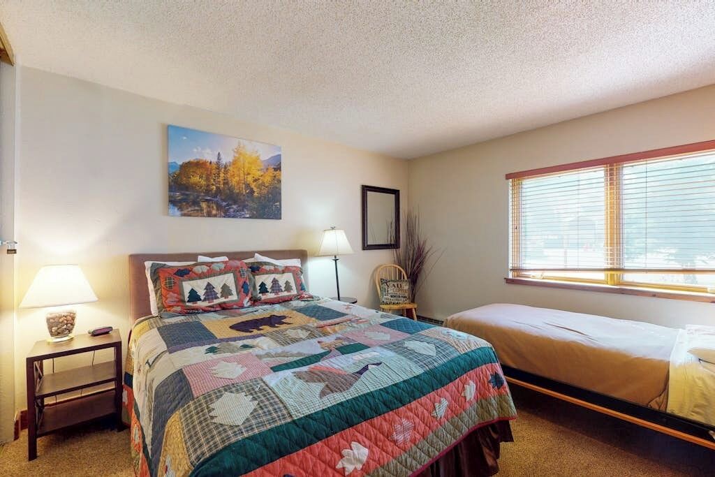 Une chambre avec un grand lit, un plus petit lit, une table de chevet avec une lampe, une chaise, une fenêtre avec des stores et un tableau au mur.