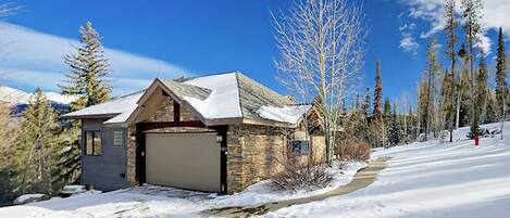 A house with a garage, surrounded by snow-covered ground and trees.
