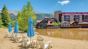 Vista de un lago con sillas de playa y sombrillas, un edificio de techo rojo y montañas al fondo.
