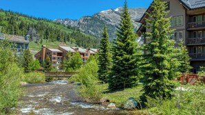 Un albergue de montaña con un río que atraviesa la propiedad, rodeado de exuberante vegetación y montañas.
