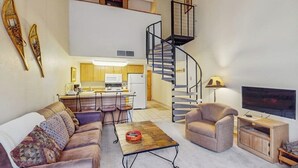 A living room with a sofa, armchair, coffee table, TV, and a spiral staircase leading to an upper level.