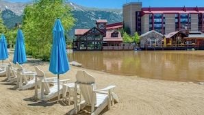 A lakeside hotel with outdoor seating, a mountain backdrop, and a modern building with red and brown accents.