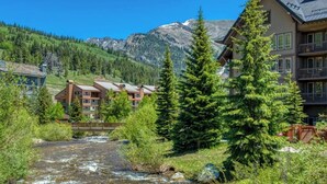 A lodge with a river in front, surrounded by mountains and greenery.