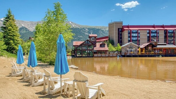 A lakeside setting with Adirondack chairs and blue umbrellas, a wooden cabin, and a modern building with a red roof. - Dog-friendly, ski condo with golf course views, balcony, hot tub & sauna (Copper Mountain)