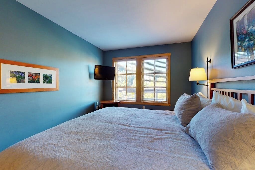 A bedroom with a bed, a TV, a window with blinds, a lamp, and framed pictures on the wall.