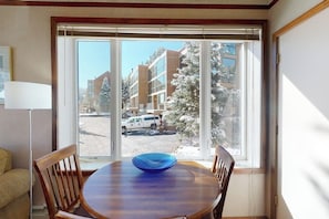 A dining area with a round table, chairs, and a view of buildings and a parked vehicle through large windows.