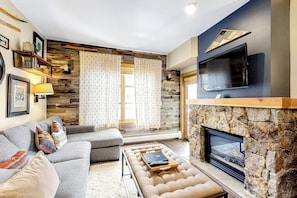 A living room with a stone fireplace, a grey sofa, a television mounted on the wall, and a window with curtains. - Spacious & Bright Condo W/ Shared Pool, Easy Ski Access & Free WiFi (Copper Mountain)