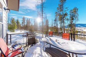 Una terraza con mobiliario exterior, una parrilla y vistas a un paisaje nevado con árboles y montañas.
