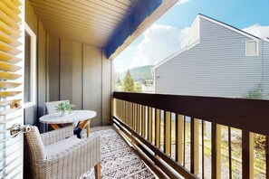 Ein überdachter Balkon mit Korbmöbeln, einem kleinen Tisch und Blick auf ein Haus und Hügel.