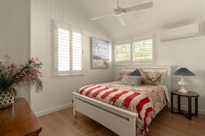 Ein Schlafzimmer mit einem Bett, einem Deckenventilator, einem Fenster mit Jalousien, einem Gemälde, einem Nachttisch und einer Lampe.