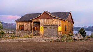 A rustic wooden house with a stone foundation, a garage, and a view of mountains and a lake. - Large mountain home with epic views, hot tub, &  gas fireplace (Grand Lake)