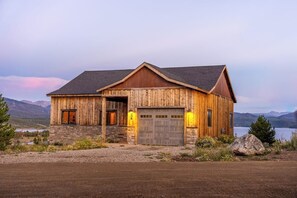 A rustic wooden house with a stone foundation, a garage, and a view of mountains and a lake. - Large mountain home with epic views, hot tub, & gas fireplace (Grand Lake)