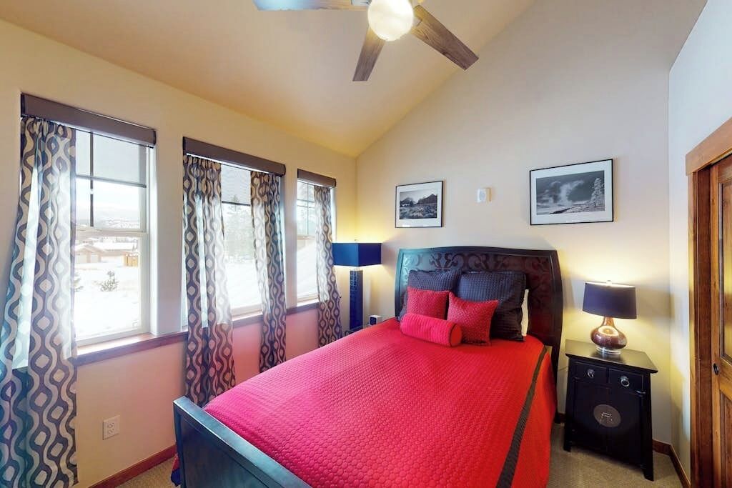 Une chambre à coucher avec un lit, des rideaux, un ventilateur de plafond, une table de chevet et deux tableaux encadrés accrochés au mur.