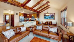 A spacious living area with a brown leather sofa set, wooden beams on the ceiling, a kitchen in the background, and a painting on the wall. - Top-floor condo with private hot tub, amazing mountain view & gas fireplace (Fraser)