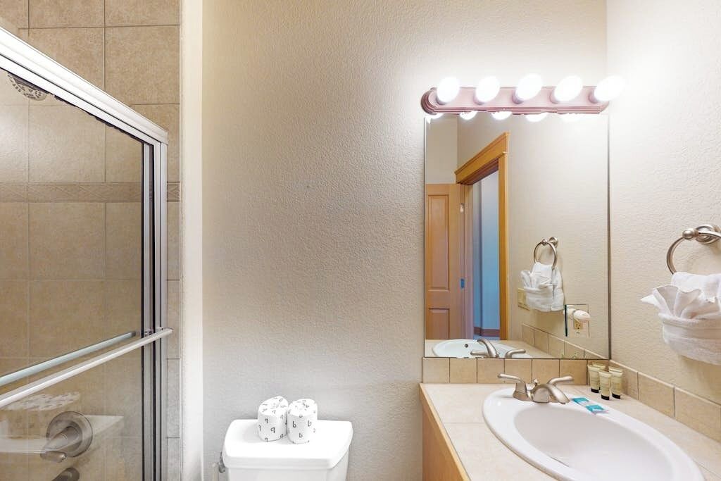 A bathroom with a shower, toilet, sink, and vanity mirror with lights.