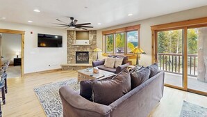 Un salón amplio con chimenea de piedra, ventilador de techo y ventanas grandes que ofrecen vistas al exterior.