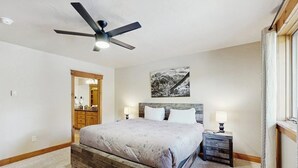 Un dormitorio con una cama, ventilador de techo, mesita de noche, lámpara de noche, cabecera de madera y un cuadro en la pared.