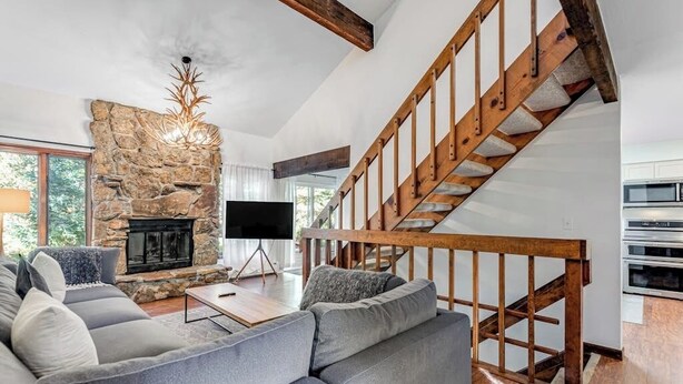 Una sala de estar moderna con chimenea de piedra, una escalera de madera, un sofá grande de color gris y un televisor de pantalla plana.