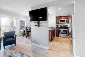 A modern kitchen with a flat-screen TV, wooden cabinets, stainless steel appliances, and a dining area with a bar stool. - Modern Waterfront Condo w/ Mountain View, W/D, & High Speed WiFi - Near Skiing (Frisco)