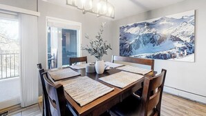 Een eethoek met een houten tafel, stoelen, een kroonluchter, een vaas met planten en een groot schilderij van een berglandschap aan de muur.