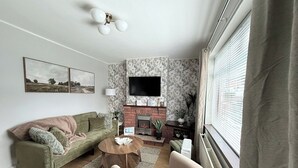 En stue med sofa, sofabord, pejs, tv ophængt på væggen og et vindue med persienner.