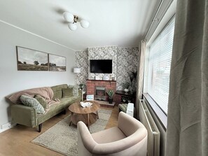 A living room with a sofa, a coffee table, a fireplace, a TV mounted on the wall, and a window with blinds. - 3BD Cozy Family Retreat for 5 (Belfast)