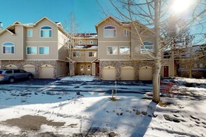 A multi-story residential building with a stone facade, multiple garage doors, and a clear driveway. - Peak-view townhome with convenient central location, decks & free EV charger (Frisco)