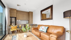 A modern living room with a leather sofa, a dining table with yellow chairs, a kitchen area, and a large mirror on the wall.
