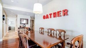 Un comedor con mesa y sillas de madera, un cartel con la palabra 'GATHER' en la pared y un pasillo que conduce a otra habitación.