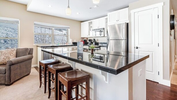 Una cocina moderna con una isla grande, armarios blancos, electrodomésticos de acero inoxidable y una zona de estar con sofá y taburetes de bar.
