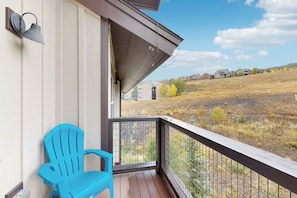 Ein Balkon mit blauem Stuhl, Metallgeländer und Blick auf ein Wiesfeld mit Gebäuden in der Ferne.
