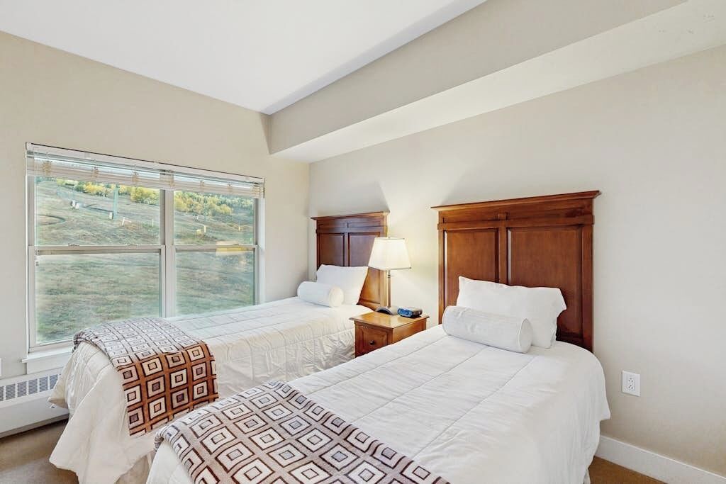 A bedroom with two beds, a window with a view, a nightstand with a lamp, and a wooden headboard.