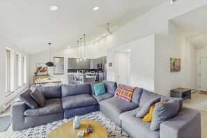 Ein modernes Wohnzimmer mit einem grauen Ecksofa, einem Holztisch und einer Küche im Hintergrund.