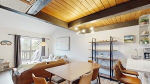 Un interno moderno con soffitto in legno, zona pranzo con tavolo e sedie, un divano e angolo cottura con scaffali e frigorifero.