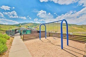 Ein Spielplatz mit Schaukeln und einem eingezäunten Bereich, ein Weg und ein Gebäude im Hintergrund.