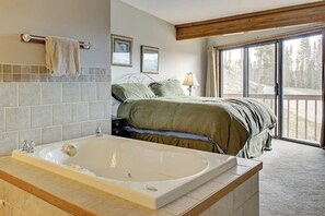 Ein Schlafzimmer mit einer großen Badewanne, einem Bett mit türkisgrüner Bettwäsche, einem Nachttisch mit Lampe und Blick durch eine Schiebetür.