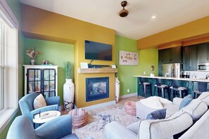 A living room with a fireplace, a television, and a kitchen area with bar stools. - Charming townhome in central locale with fireplace & balcony - dog-friendly (Granby)