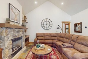 Ein Wohnzimmer mit Kamin aus Stein, einem großen braunen Ecksofa, einem runden Couchtisch mit Dekoration, einer Wanduhr und einem gerahmten Bild.