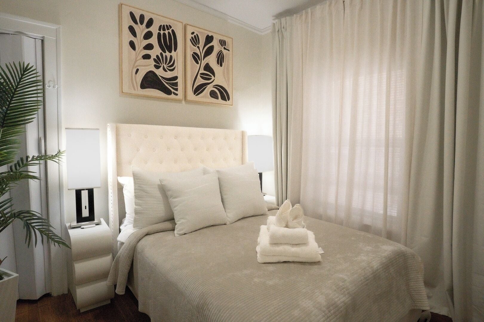 A hotel room with a bed, pillows, a bedside table, a lamp, a plant, and artwork on the wall.