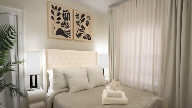 A hotel room with a bed, pillows, a bedside table, a lamp, a plant, and artwork on the wall. - 6BR Apt • Workspace • Near Lakefront + Red Line (Chicago)