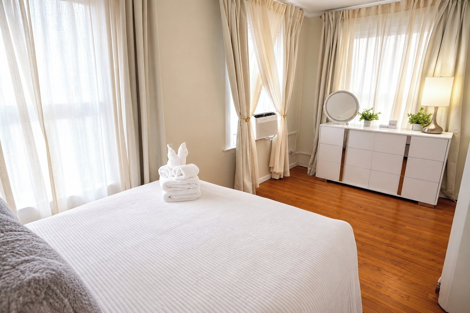A hotel room with a neatly made bed, a dresser with a mirror, and a bedside table with a lamp.