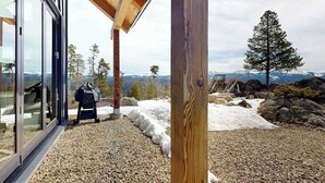 Un edificio moderno con amplios ventanales de vidrio, una terraza de madera y vistas a terreno cubierto de nieve y montañas distantes.